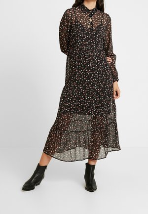 Frau trägt ein schwarzes, durchsichtiges Midikleid mit Blumenmuster, langen Ärmeln, gestufter Rock, mehrere Halsketten und schwarze Knöchelstiefel, steht auf weißem Boden.