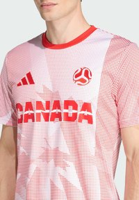 Punainen ja valkoinen lyhythihainen jalkapallopaita, jossa on pistekuvio, esittelee "KANADA"-tekstin selvästi punaisella, Adidas-logo vasemmassa olkapäässä.