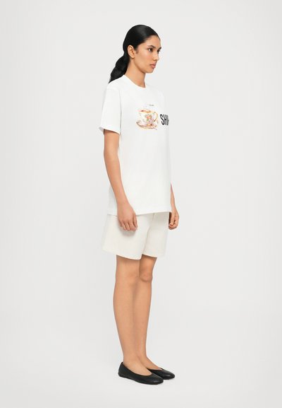 Tricou alb cu grafică de cană de ceai florală și text "SHIO", asortat cu pantaloni scurți bej deschis și pantofi slip-on negri. Textura materialului este netedă.