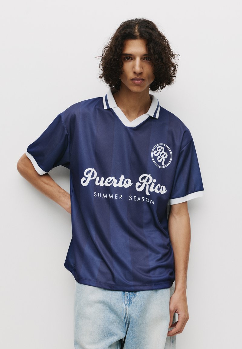 T-shirt à manches courtes bleu marine avec des accents blancs, comportant "Puerto Rico" et "SAISON D'ÉTÉ" imprimés en lettres blanches. Design en V.