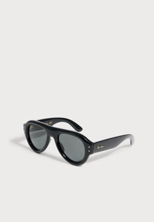 Ochelari de soare Ray-Ban negri cu lentile rotunjite și logo auriu pe braț, așezați pe o suprafață gri deschis.