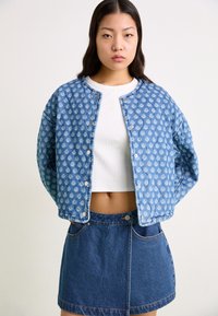 Μπλε επενδυμένο μπουφάν με λουλουδάτο σχέδιο και ασημένια κουμπιά πίεσης, σε συνδυασμό με μια τζιν φούστα. Και τα δύο ρούχα διαθέτουν χαλαρό σχέδιο.