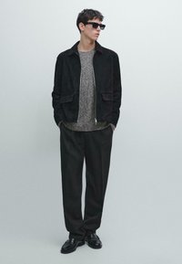 Veste en suede noire, pull gris texturé, pantalon noir ample et chaussures en cuir noires, avec des lunettes de soleil carrées. Le mannequin se tient devant un arrière-plan clair.