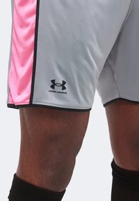 Κοντινή λήψη γκρι και ροζ αθλητικών σορτς Under Armour που φοράει ένα άτομο με σκούρο δέρμα και μαύρες κάλτσες μέχρι το γόνατο.
