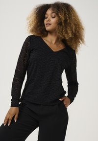 Top in pizzo nero con maniche lunghe e scollatura a V, caratterizzato da un motivo floreale e una finitura testurizzata. Abbinato a pantaloni neri.