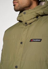 Zatvoren kadar muškarca u maslinastozelenoj Berghaus jastučić jakni s visokim ovratnikom i crnim kopčama na dugmad.