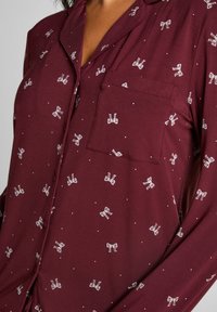 Bordeauxrode pyjamabovenkant met lange mouwen, gemaakt van zachte stof, met witte strikpatronen en kleine stippen, voorzien van een borstzakje en knoopsluiting.