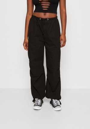 Person trägt schwarze Hosen mit weitem Bein und Kordelzugbündchen, ein schwarzes Crop-Top mit Ausschnitten und schwarz-weiße Sneaker, steht auf einfachem Boden.