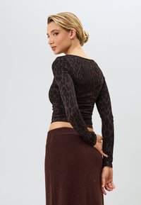Langärmliges Oberteil in Dunkelbraun mit schwarzem Leopardenmuster, figurbetonter Schnitt und gerüschter Textur. Getragen zu einem braunen Strickrock, von hinten betrachtet.