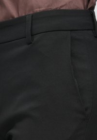 Pantalon noir avec une texture de tissu lisse, présentant un devant plat, des passants de ceinture et des poches latérales, conçu pour une coupe ajustée.