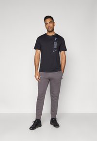 Schwarzes Baumwoll-T-Shirt mit blauem Nike-Logo und Druck, kombiniert mit grauen Trainingshosen mit Nike-Akzent, getragen mit schwarzen Sportschuhen.