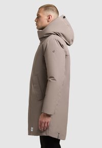 Bézs színű szigetelt parka magas gallérral és nagy kapucnival, oldalzsebekkel, elegáns kialakítással és egy márkás címkével az alsó szegélyen.