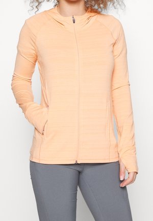 Veste zippée orange clair à capuche, dotée de manches longues, d'un tissu texturé et de poches latérales. Portée avec un pantalon gris.