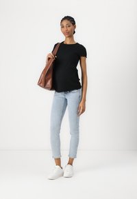 Camiseta negra de rayas, jeans azul claro, zapatillas blancas. Sostenido un bolso de cuero marrón en el hombro, de pie contra un fondo blanco sencillo.