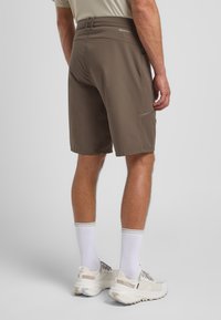 Pantaloni corti marroni realizzati in materiale leggero, con vestibilità slim, tasca laterale e logo in vita. Indossati con calzini bianchi alla caviglia e scarpe.
