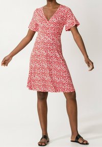 Robe fleurie rouge avec un décolleté en V, des manches courtes et un ourlet évasé. Un motif de fleurs blanches recouvre le tissu. Portée avec des sandales plates noires.