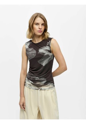 Femme aux cheveux blonds mi-longs portant un haut noir sans manches avec un grand imprimé floral blanc et un pantalon ample couleur crème, debout devant un fond blanc.