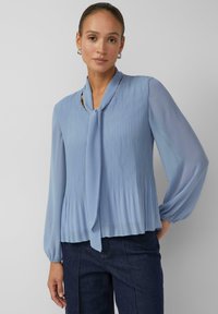 Hellblaue Bluse mit plissiertem Design, langen Puffärmeln, Binddetail am Ausschnitt und weichem, fließendem Stoff. Kombiniert mit dunklen Jeans.