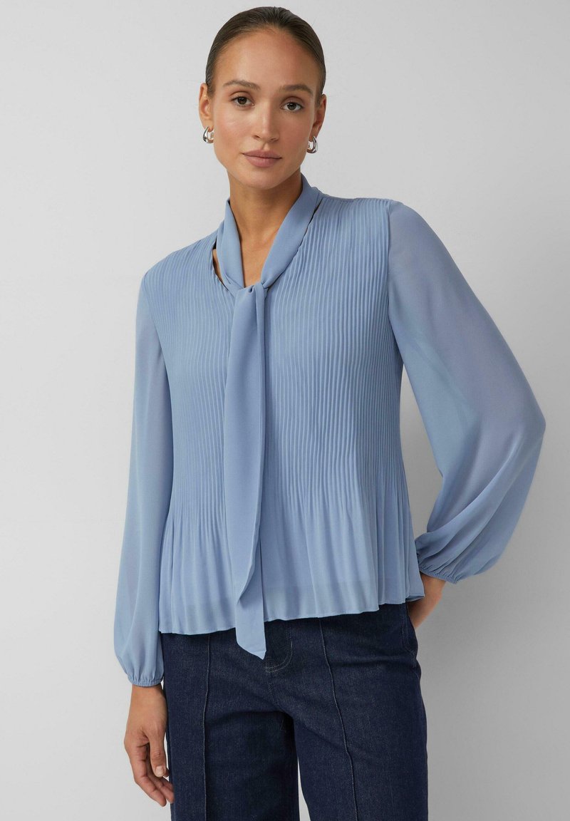 Hellblaue Bluse mit plissiertem Design, langen Puffärmeln, Binddetail am Ausschnitt und weichem, fließendem Stoff. Kombiniert mit dunklen Jeans.