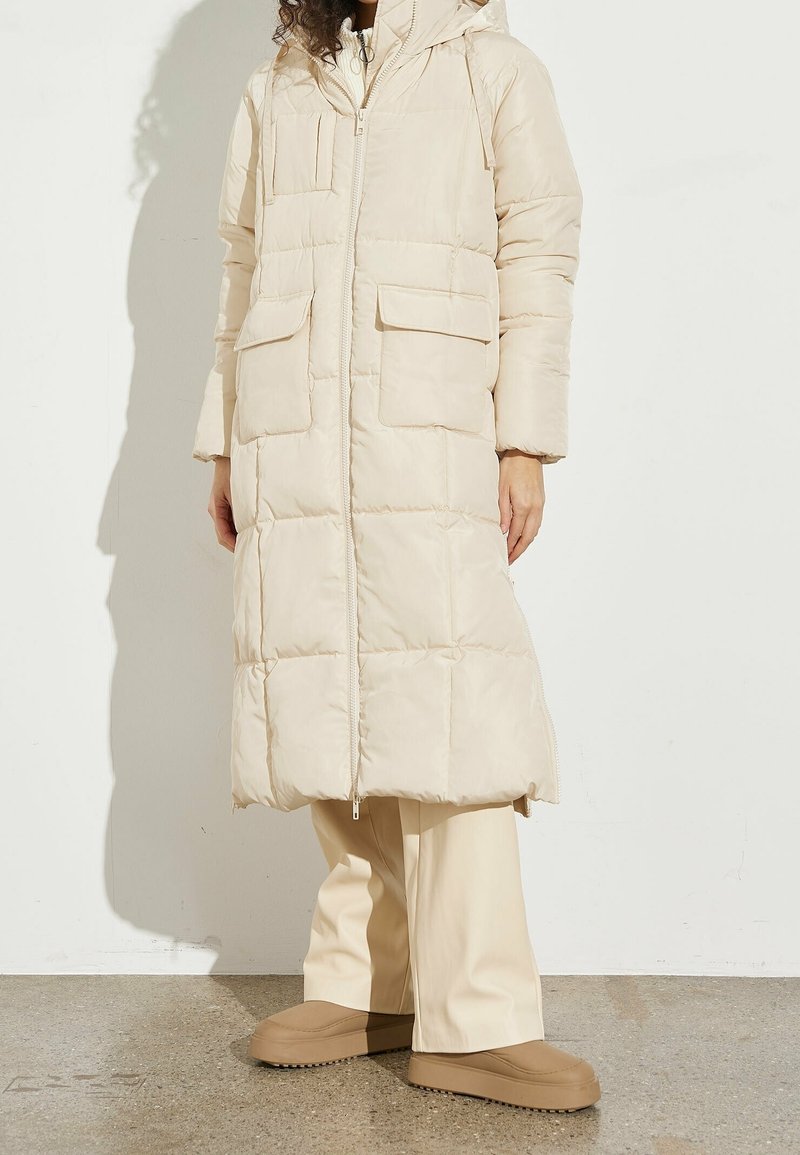 Femme portant un long manteau matelassé beige avec capuche, pantalon crème à jambes larges, et chaussures beige à enfiler, debout sur un sol en pierre.