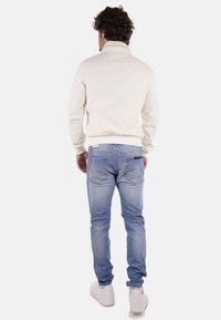 Giacca bomber crema sopra jeans azzurri chiari. La giacca presenta una texture liscia, polsini a costine e un colletto alto. Sneakers bianche completano il look.