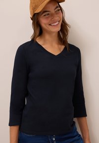 Marineblaues, langärmeliges V-Ausschnitt-Oberteil aus weichem Stoff, kombiniert mit einer beige Kappe; hat einen lässigen Schnitt und minimale Nähdetails.