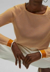 Gestreifter Pullover in Orange und Weiß, mit einem runden Halsausschnitt, gerippten Bündchen und einem strukturierten Strickdesign. Die Hände sind in die Hüften gestützt.