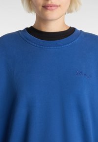 Modrá mikina z jemné bavlny, s žebrovaným kulatým výstřihem a decentně vyšitým logem na hrudi. Hladký materiál, ležérní design.