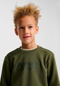 Ung pojke med lockigt blont hår, iklädd en olivgrön Hummel Sportswear-tröja, som tittar något åt sidan mot en enfärgad bakgrund.