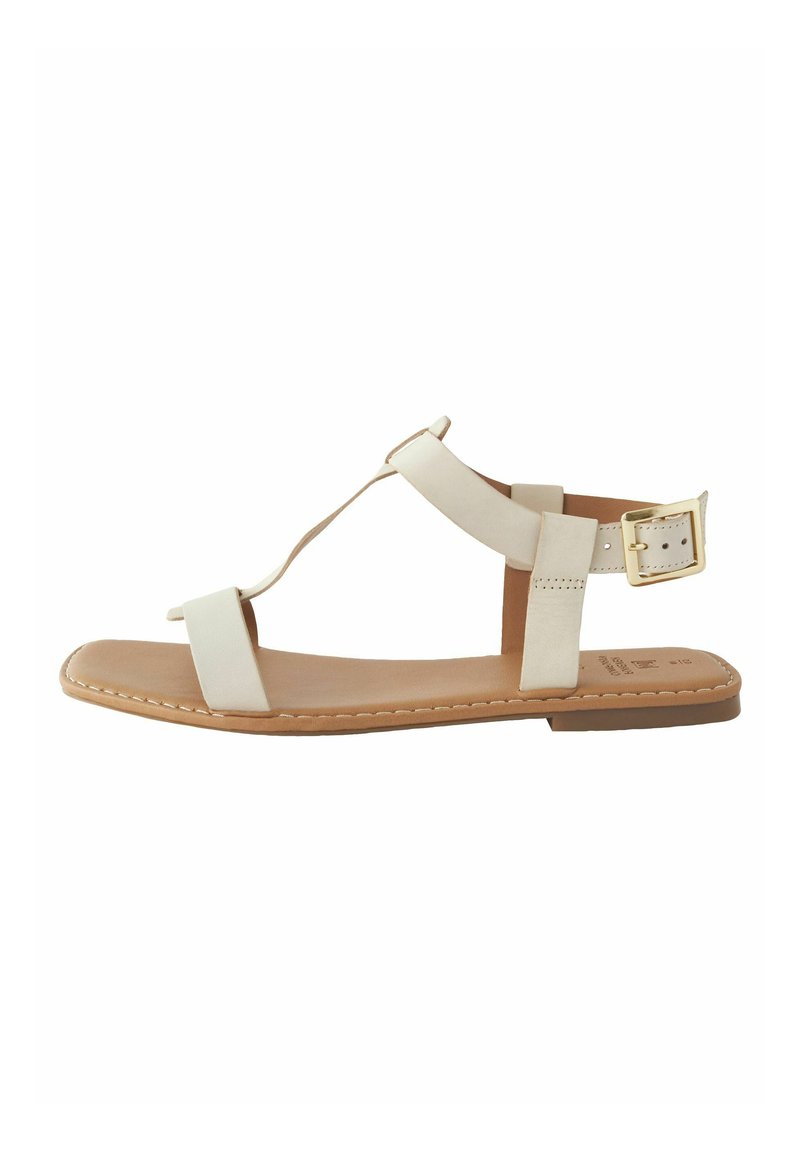 Vit platt sandal med T-rem design, justerbar vristrem, mjukt lädermaterial och en ljusbrun sula. Guldspänne som detalj.