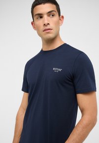 Granatowy bawełniany t-shirt z krótkim rękawem, z okrągłym dekoltem i małym białym logo "MUSTANG" na piersi. Gładka tekstura.