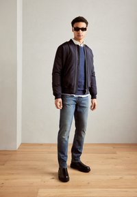 Navy bomber jacket over a blue sweater, white shirt, and blue jeans. Black shoes with thick soles. Short, curly hair. Sunglasses worn.