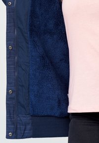 Marinblå kavaj med ett mjukt, fuzzy inre foder. Det yttre materialet är slätt med subtila ränder. Ribbad mudd och nederkant i kontrasterande textur.