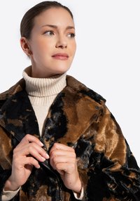 Kobieta o jasnej karnacji i włosach związanych w kok nosi beżowy sweter z golfem oraz brązowo-czarny płaszcz z sztucznego futra, zapinając zamek.