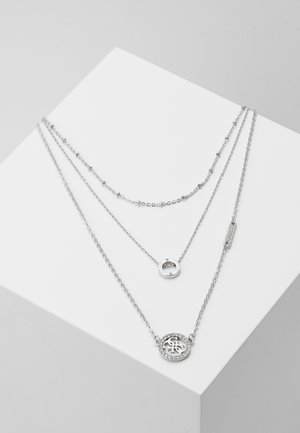 Drie gelaagde zilveren kettingen met delicate schakels; één met kleine kralen, één met een ronde hanger en één met een ronde logohanger.