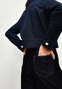 Chaqueta de mezclilla oscura con un diseño corto, que presenta un cuello puntiagudo y puños abotonados. Botones dorados destacados y detalles de costura en contraste.