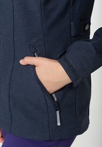 Giacca blu navy con tasca con zip, tessuto testurizzato e dettagli in velcro sui polsini. Primo piano di una mano nella tasca.