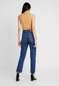 Bruine geribde tanktop met blauwe high-waisted denim jeans. Opvallende donkere stiksels en zakdetails. Draag met zwarte hoge hakken.