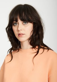 Sweat-shirt de couleur pêche avec un col rond et des manches courtes, arborant un logo discret et une texture de tissu lisse et léger.