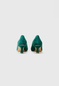 Un paio di scarpe in suede verde acqua con tacco kitten viste dal retro su uno sfondo bianco semplice.