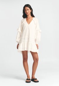 Valge puuvillane kleit V-kaelusega, volangvarrukatega ja kihilise hemiga. Kanga pinnal on tekstureeritud viimistlus. Kandmisel mustade sandaalidega.