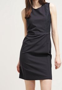 Svart ärmlös klänning med en figurnära silhuett. Har ett spetspanel på ena sidan och slät tyg. Knälång med en lätt lyster.