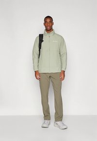 Chaqueta verde claro de material ligero, con cremallera completa al frente, combinada con pantalones caqui y zapatillas blancas. Mochila negra sobre el hombro izquierdo.