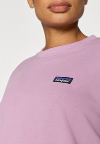 Lys lilla Patagonia-genser med rund hals og en liten merke-patch med svarte og blå fjellmotiver. Glatt tekstur.