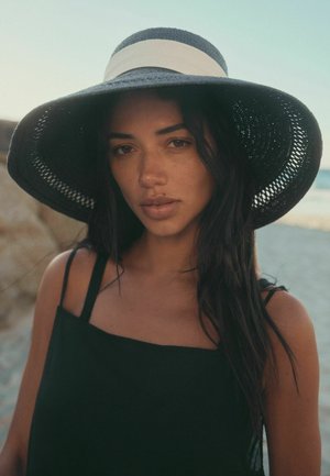 Frau mit langen dunklen Haaren, die einen breitkrempigen schwarz-weißen Hut und ein schwarzes ärmelloses Oberteil trägt, steht im Freien in der Nähe eines Strandes.