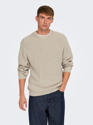 Giovane uomo con capelli corti che indossa un maglione beige a maglia e jeans blu scuro, in piedi con una mano nella tasca davanti a uno sfondo semplice.