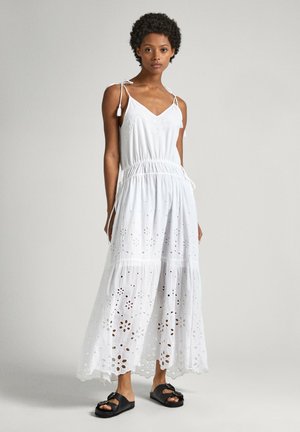 Abito maxi in cotone bianco con ricami ad occhiello, spalline regolabili, vita elastica e gonna a strati indossato da un modello con sandali neri.