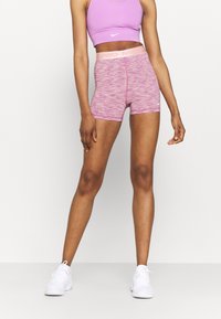 Kobieta stoi w różowym staniku sportowym Nike oraz różowych szortach Nike Pro w wzory, w białych sneakersach, na jednolitym tle.