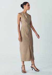 Vestido sin mangas color caqui con cremallera frontal, bolsillos laterales y dobladillo a media longitud. Combinado con tacones negros  con tiras. Textura suave.