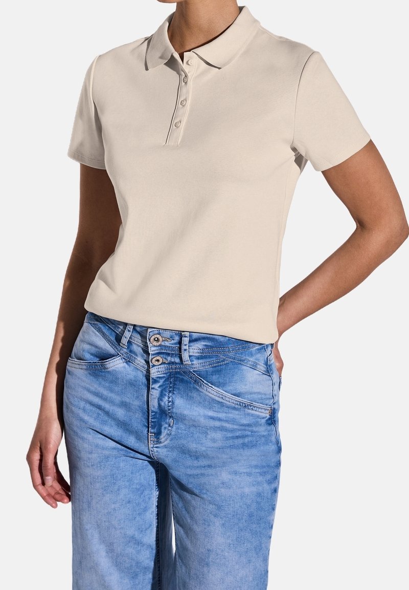Polo beige clair avec un col classique, patte de boutonnage à quatre boutons et manches courtes, associé à un jean en denim bleu clair avec un léger délavage.